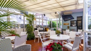 Un patio iluminado por el sol con mobiliario de mimbre blanco, plantas en macetas y un letrero que dice 'Hotel Elcano'.