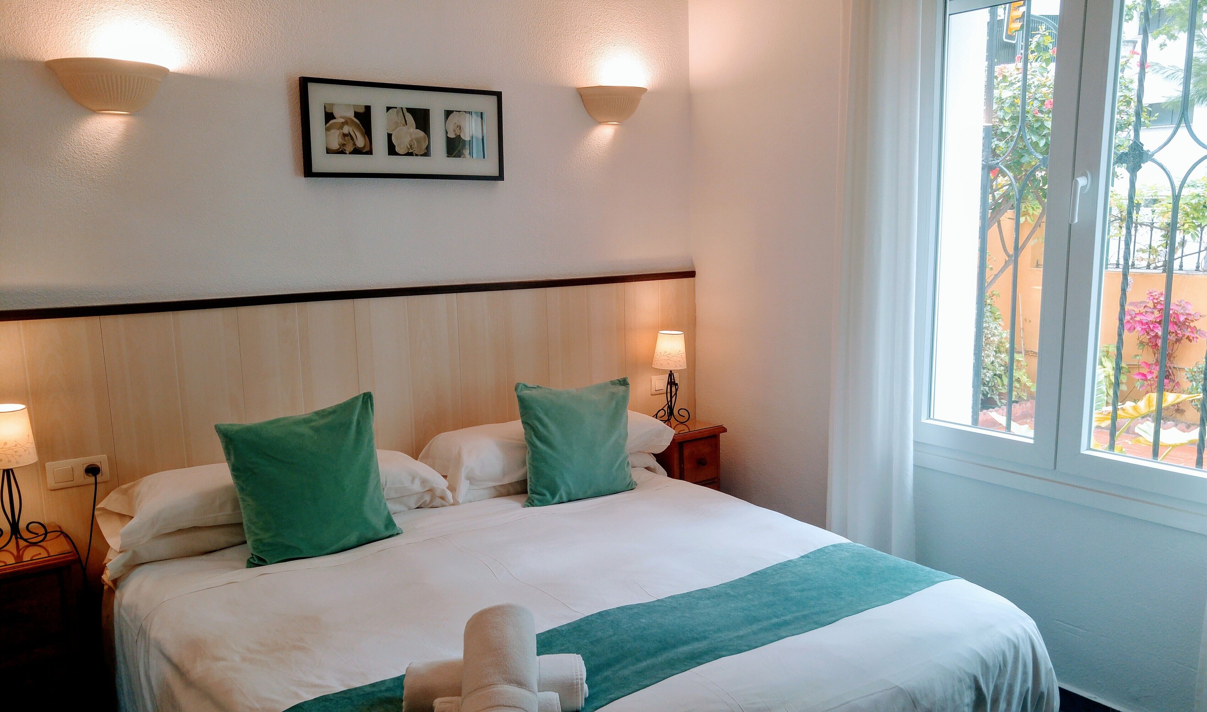 Een net opgemaakt bed met groene kussens, een bedlampje en een ingelijst schilderij aan de muur.