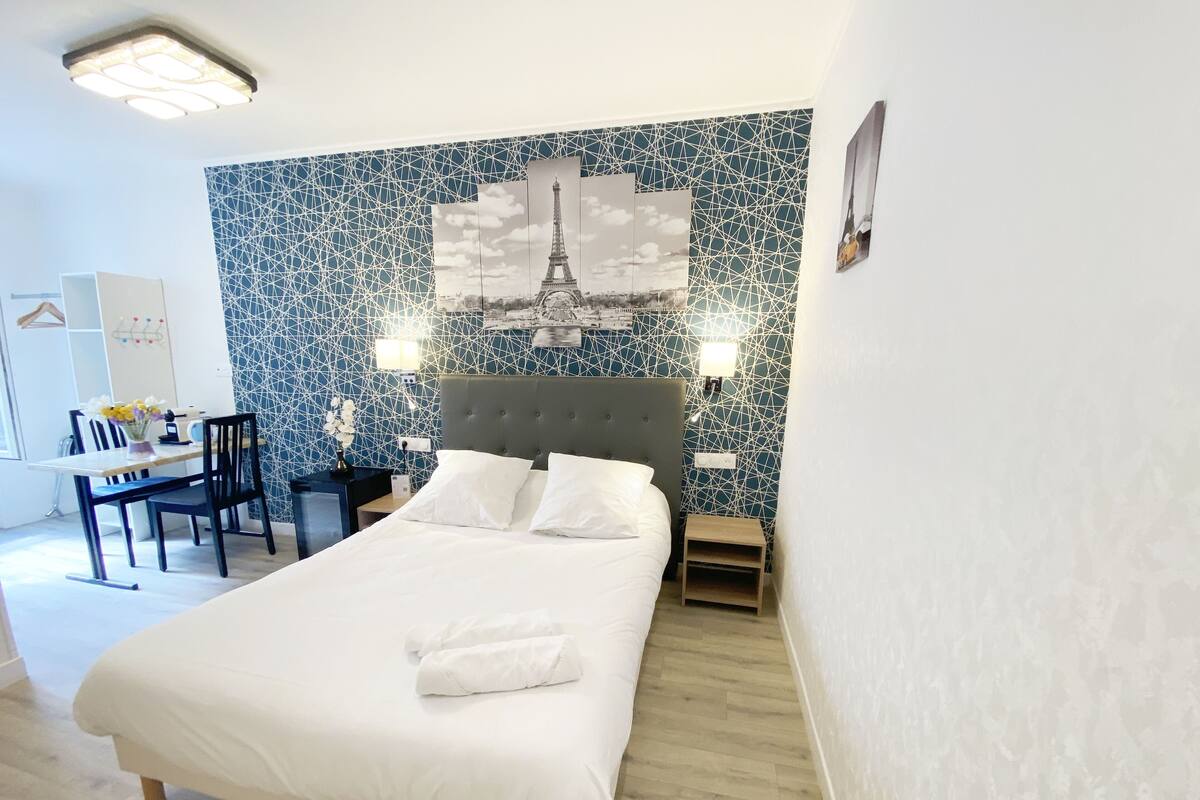 Una camera d'albergo con un letto, comodini, una scrivania con sedie, e una parete con un motivo geometrico e una foto della Torre Eiffel.