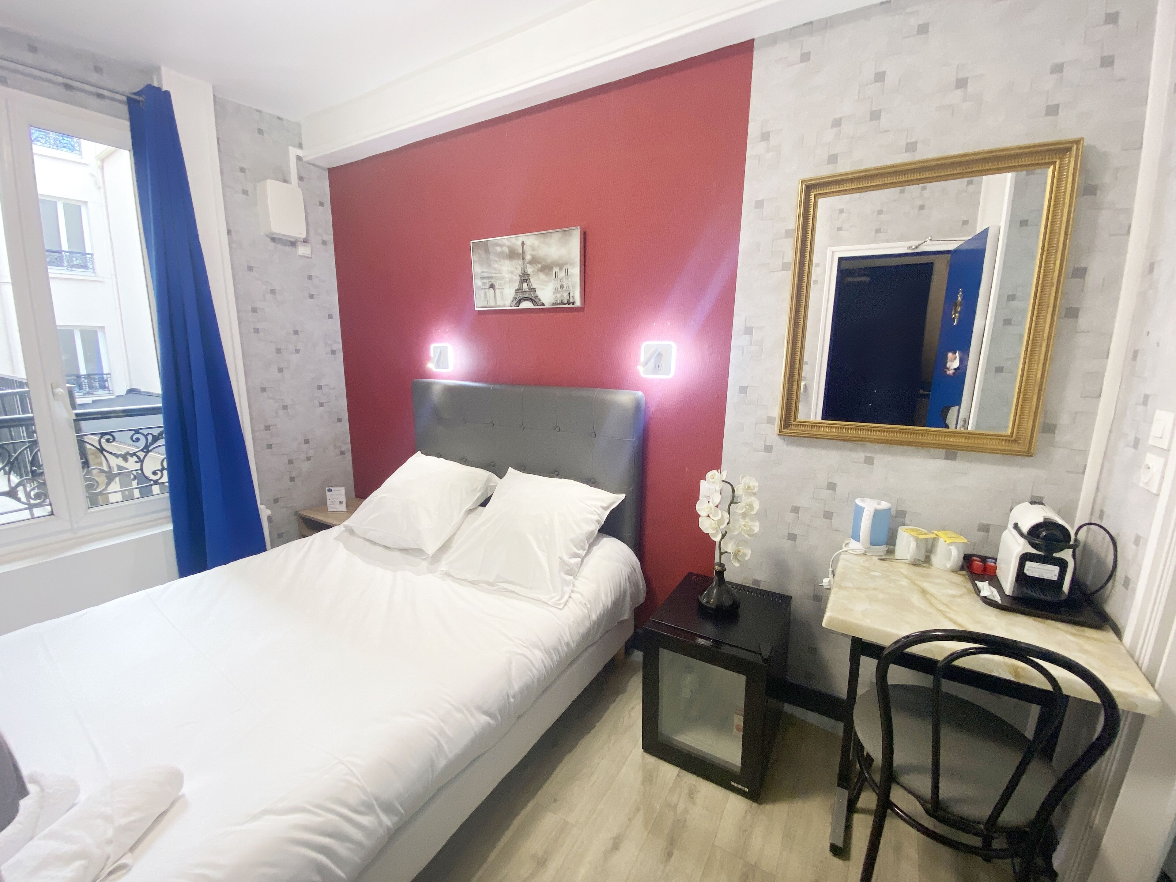 A hotel room with a bed, a nightstand, a desk with a chair, a mirror, and a window with curtains.