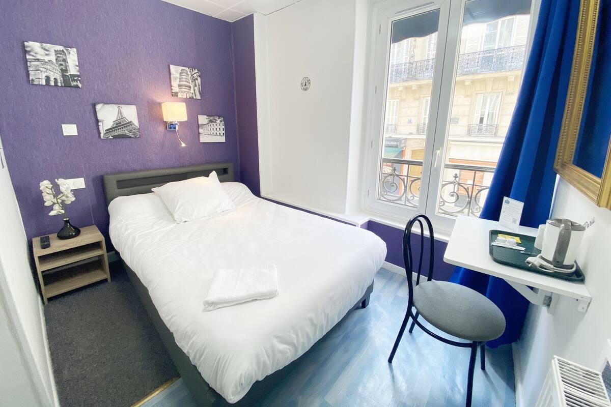 Una camera d'albergo con un letto, una scrivania con una sedia, una finestra che dà su edifici e opere d'arte alle pareti.