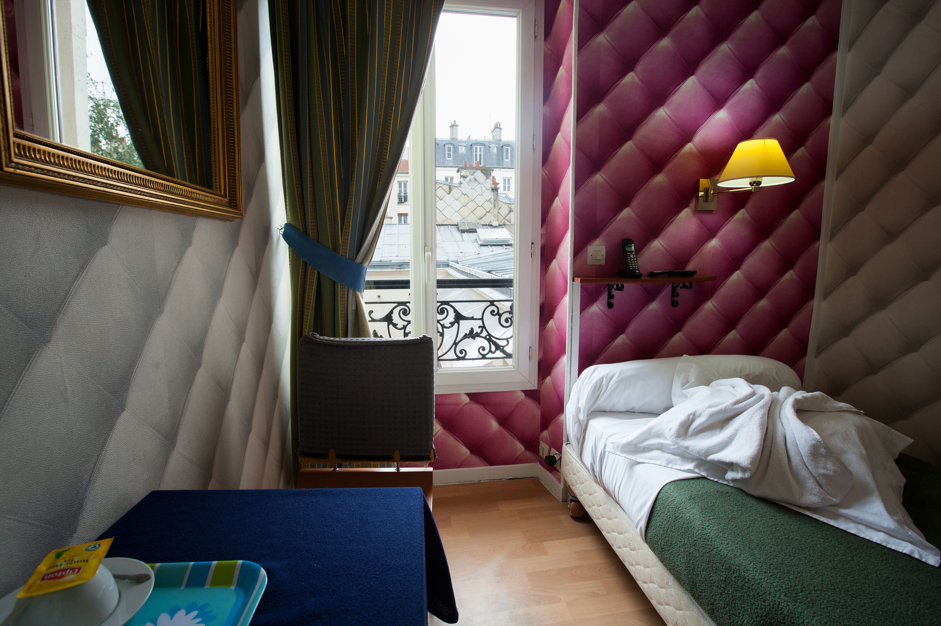 Une chambre à coucher avec un lit, une fenêtre avec des rideaux, une lampe de chevet et une vue sur les bâtiments à l’extérieur.