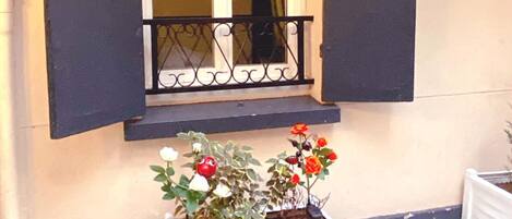 Una ventana con contraventanas oscuras, una jardinera blanca con plantas y una pequeña cesta de metal negra sobre el suelo.