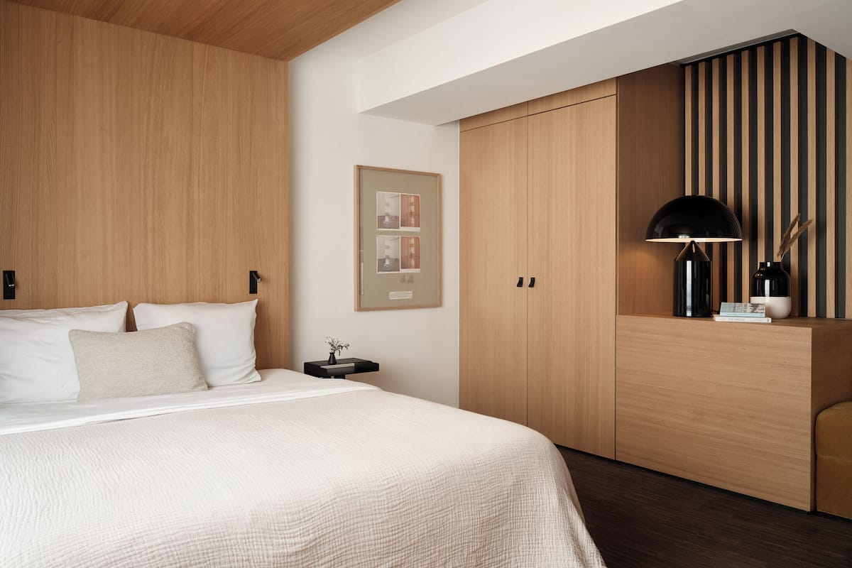 Un dormitorio moderno con techo de madera, una cama grande con ropa blanca, un armario de madera y una mesita de noche con una lámpara y un pequeño florero.