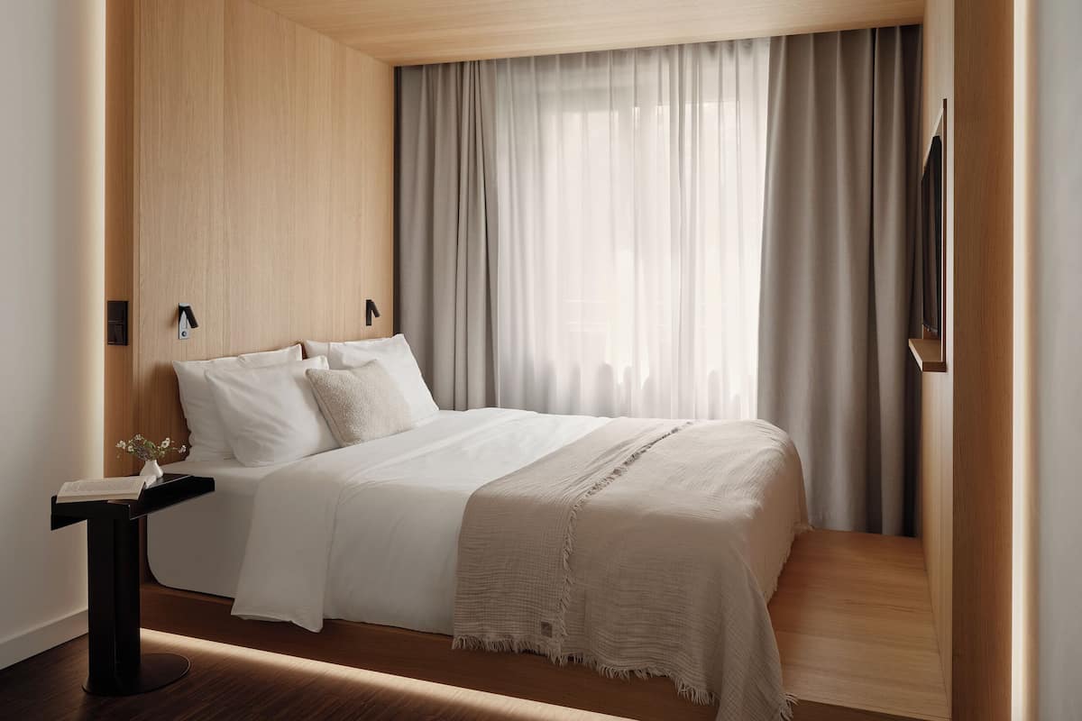 Un dormitorio moderno con cabecera de madera, una mesita de noche con un libro y un ventanal con cortinas.