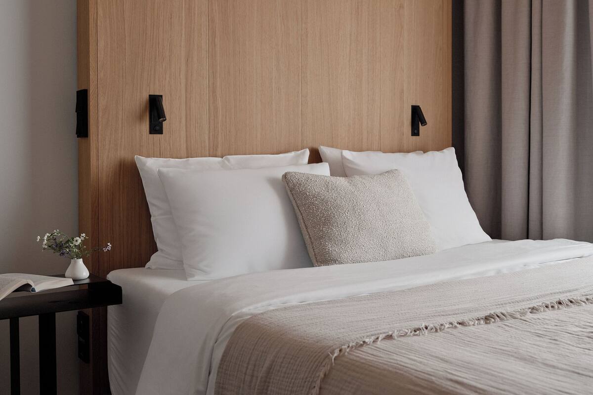 Una cama bien tendida con sábanas blancas y un plaid beige, una mesita de noche con un florero y un cabecero de madera con dos lámparas fijadas a la pared.