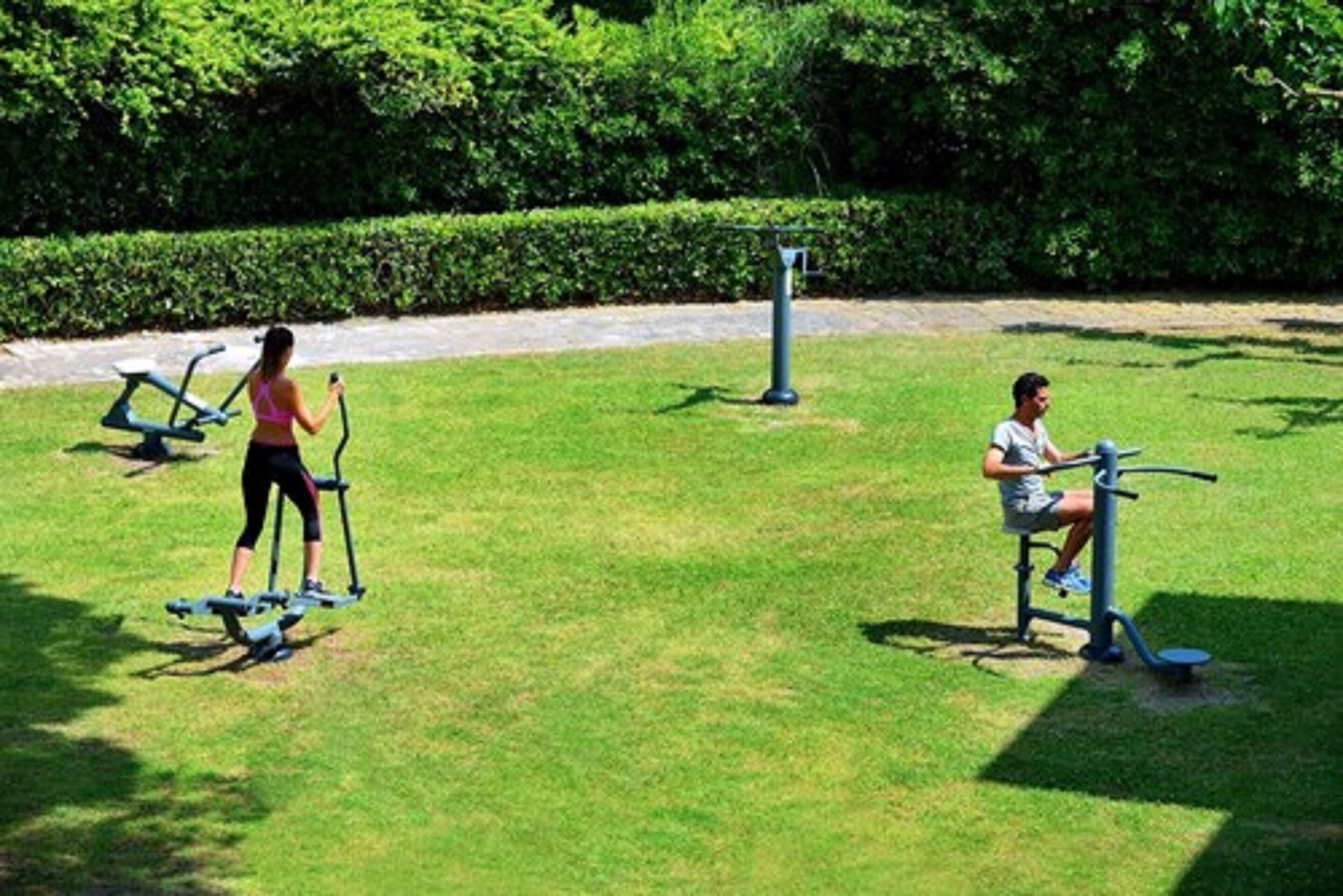 Two people exercising on outdoor fitness equipment in a park with a hedge in the background.
