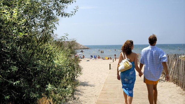 A couple walking on a wooden boardwalk beside a beach with people swimming in the water. - Residéal La Grande Motte (La Grande-Motte)