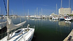 A marina with multiple sailboats docked, a large building in the background, and calm water. - Residéal La Grande Motte (La Grande-Motte)