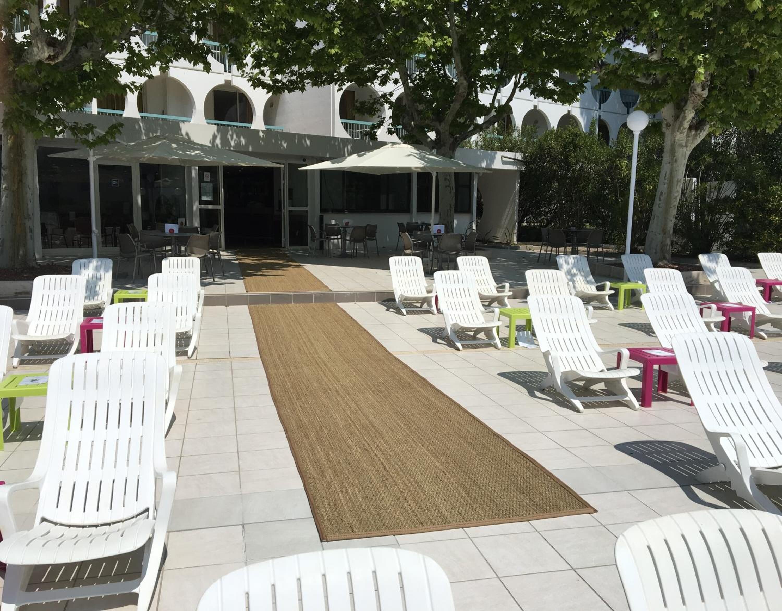 An outdoor seating area with white plastic chairs and tables, a brown mat, and a building with arched windows in the background.