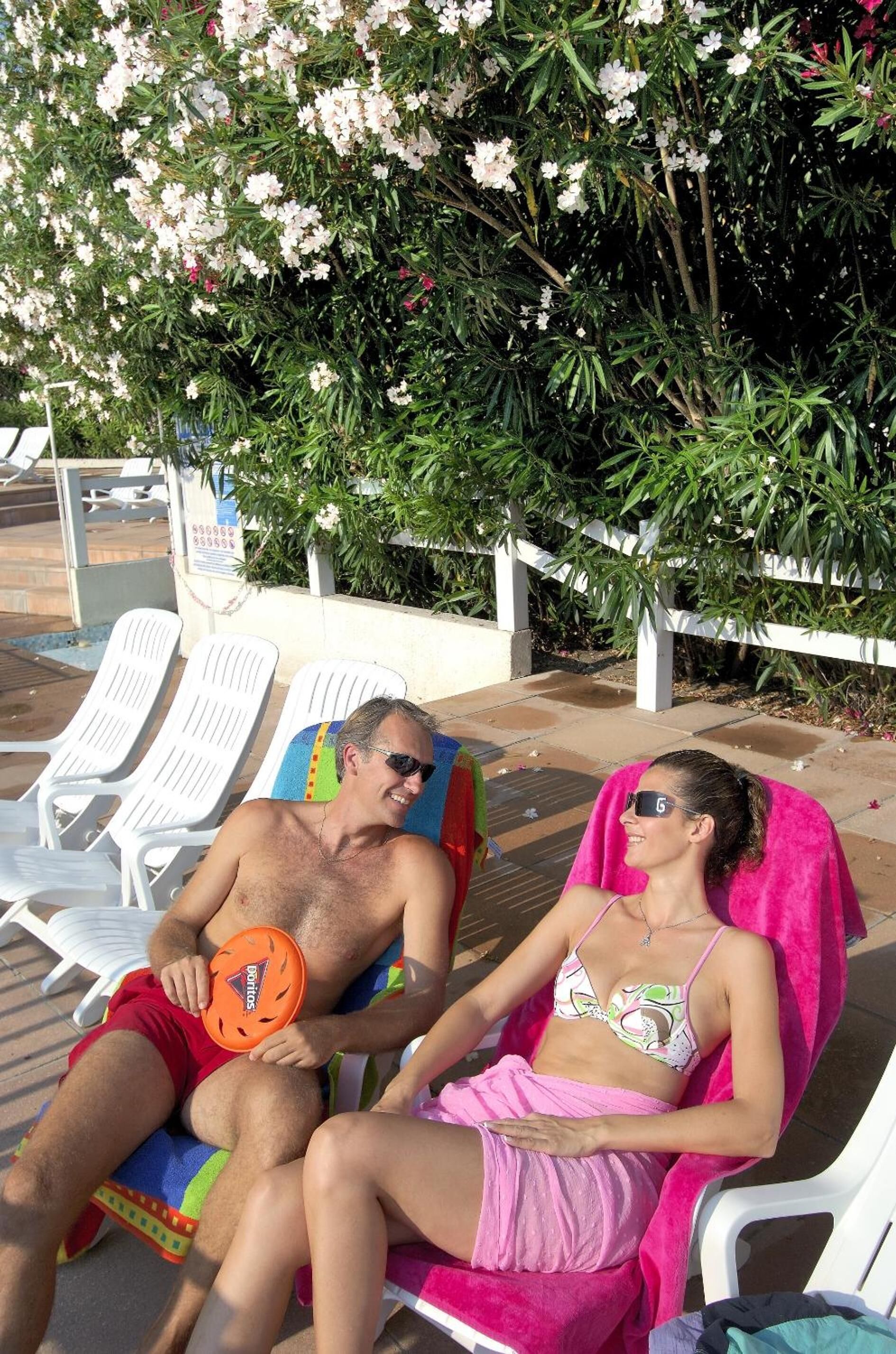 Dos personas descansando en tumbonas con una piscina y un árbol al fondo.