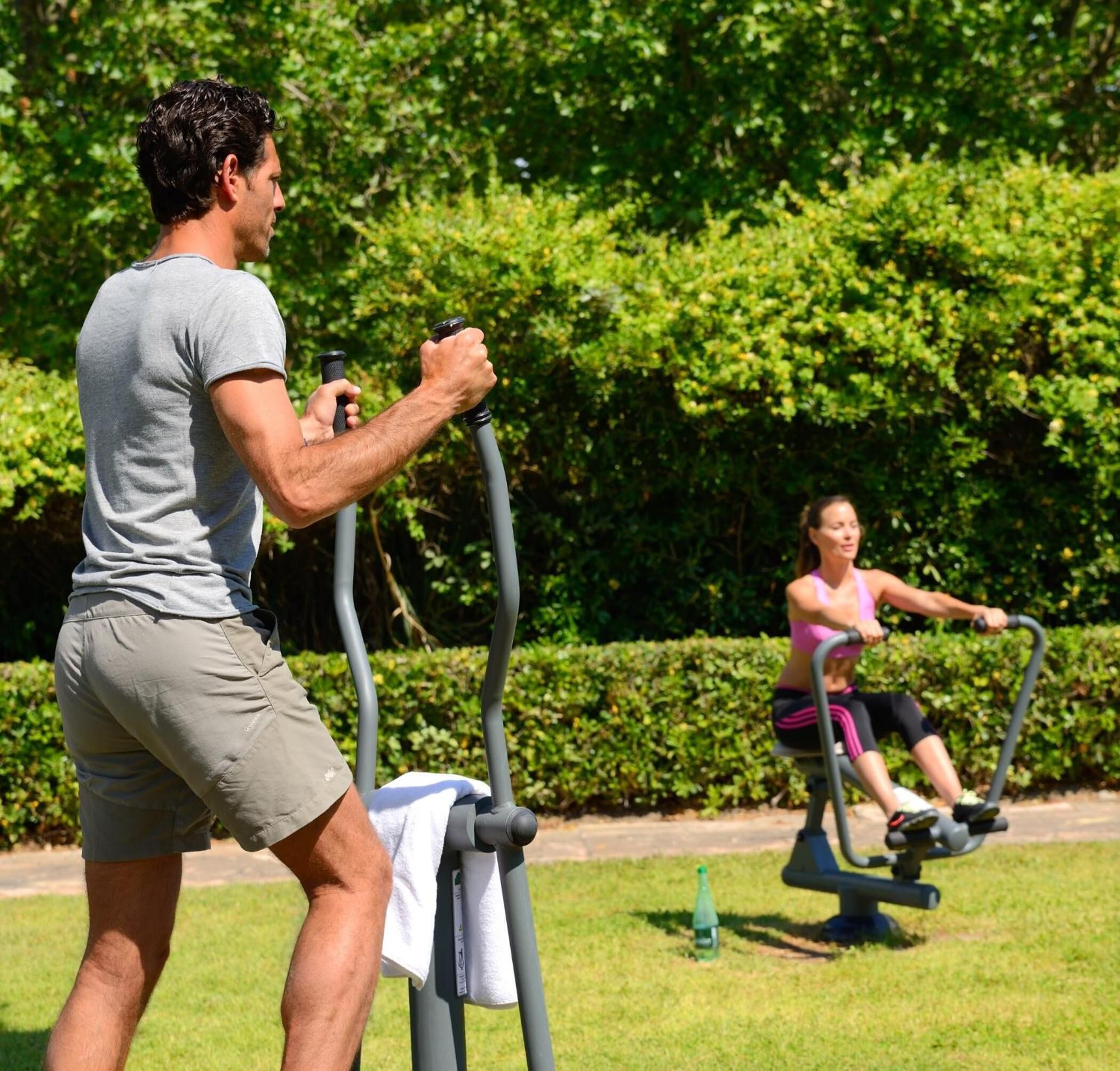 Dos personas ejercitándose en bicicletas estáticas al aire libre en un parque con césped verde y árboles.