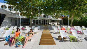 A poolside area with sun loungers, a wooden deck, and a building with arched windows in the background. - Residéal La Grande Motte (La Grande-Motte)