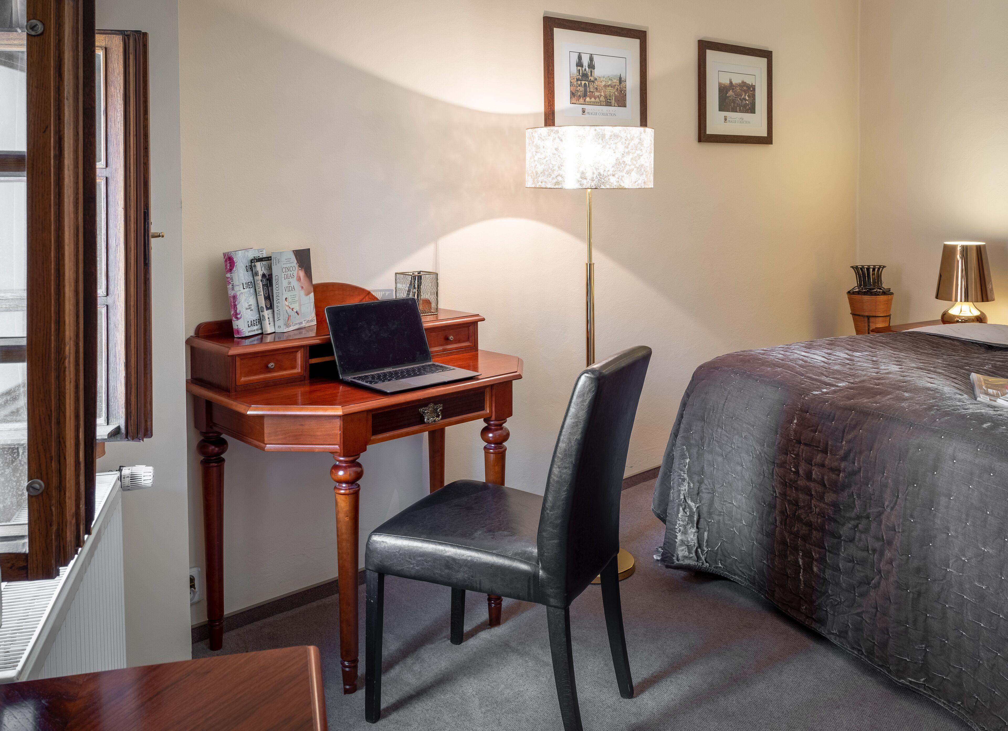 A hotel room with a wooden desk, a laptop, a chair, a bed with a dark cover, a floor lamp, and two framed pictures on the wall.