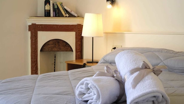 A neatly made bed with rolled towels, a bedside lamp, and a bookshelf with books and a bottle. - Residenza Le Rondini (Florence)