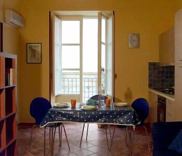 Un área de comedor con una mesa puesta para dos, una estantería y una cocina visible a través de un umbral abierto.
