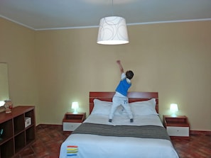 Un enfant saute sur un lit dans une chambre d’hôtel, où l’on aperçoit deux tables de chevet, une commode et une lampe de chaque côté du lit.
