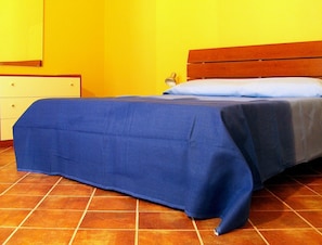 A bed with a blue bedspread, a wooden headboard, and a white pillow against a yellow wall. - Politi Aparthotel (Catania)