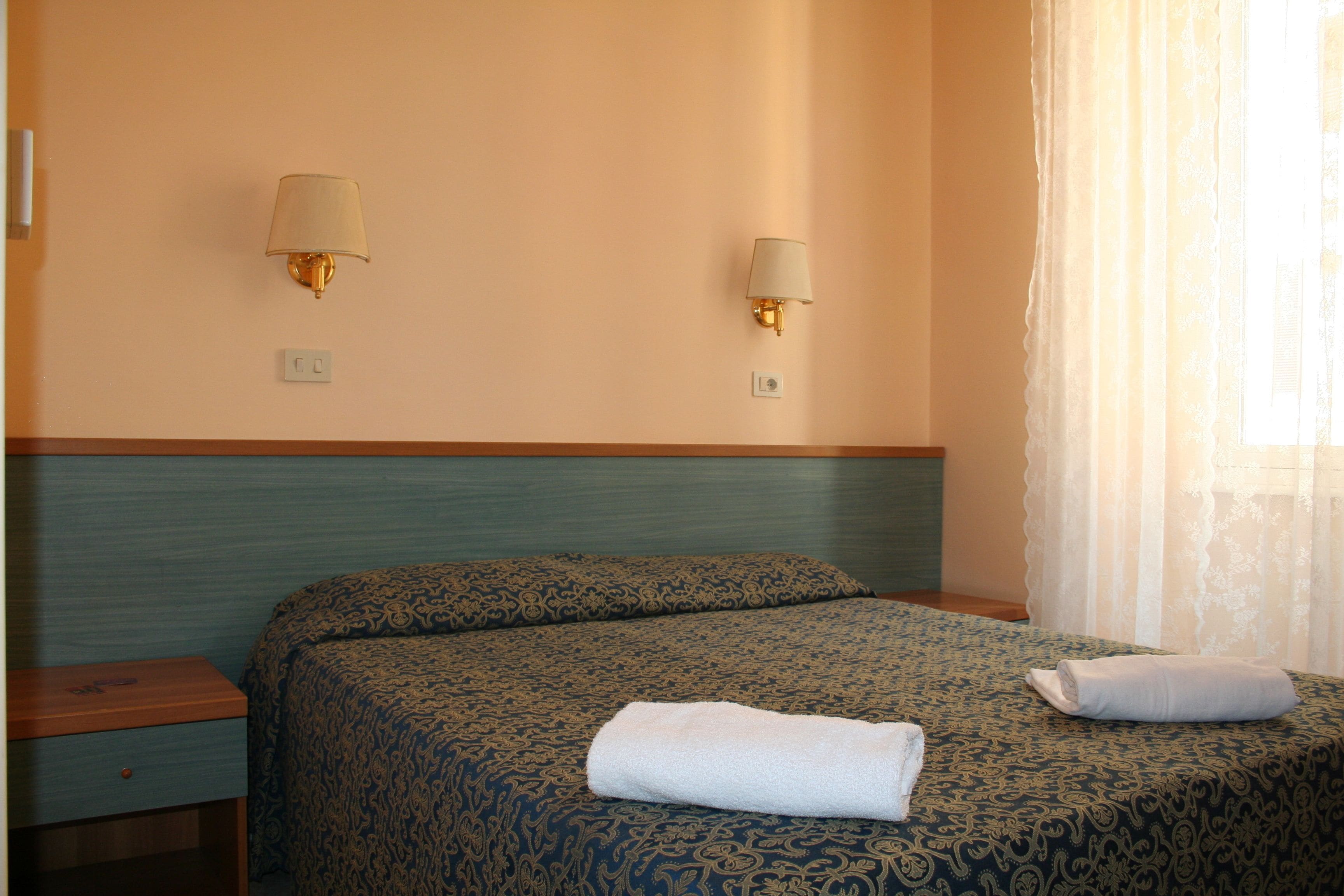 A hotel room with a bed, two wall-mounted lamps, a nightstand, and a window with sheer curtains.