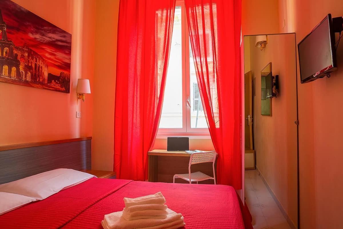 Ein Hotelzimmer mit Bett, Schreibtisch mit Laptop, Stuhl, Fernseher und einem Bild an der Wand.