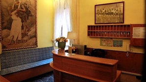 A reception area with a wooden desk, a painting on the wall, a window with curtains, and a shelf with framed photos. - Castelfidardo (Rome)