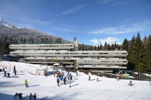 Um edifício de vários andares com um teleférico ao fundo, cercado por árvores cobertas de neve e um céu claro.