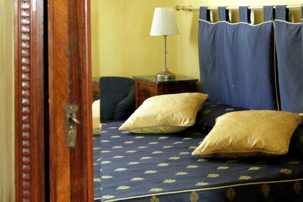 A bedroom with a wooden door, a bed with blue and gold bedding, a nightstand with a lamp, and a chair.