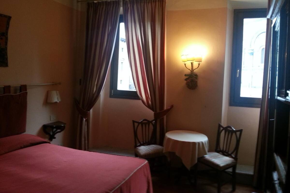 A hotel room with a bed, two chairs, a small table, a lamp, and a window with curtains.