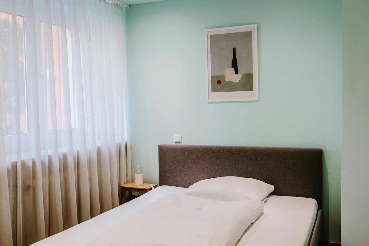 A hotel room with a bed, a nightstand, a framed picture on the wall, and sheer curtains on the window.