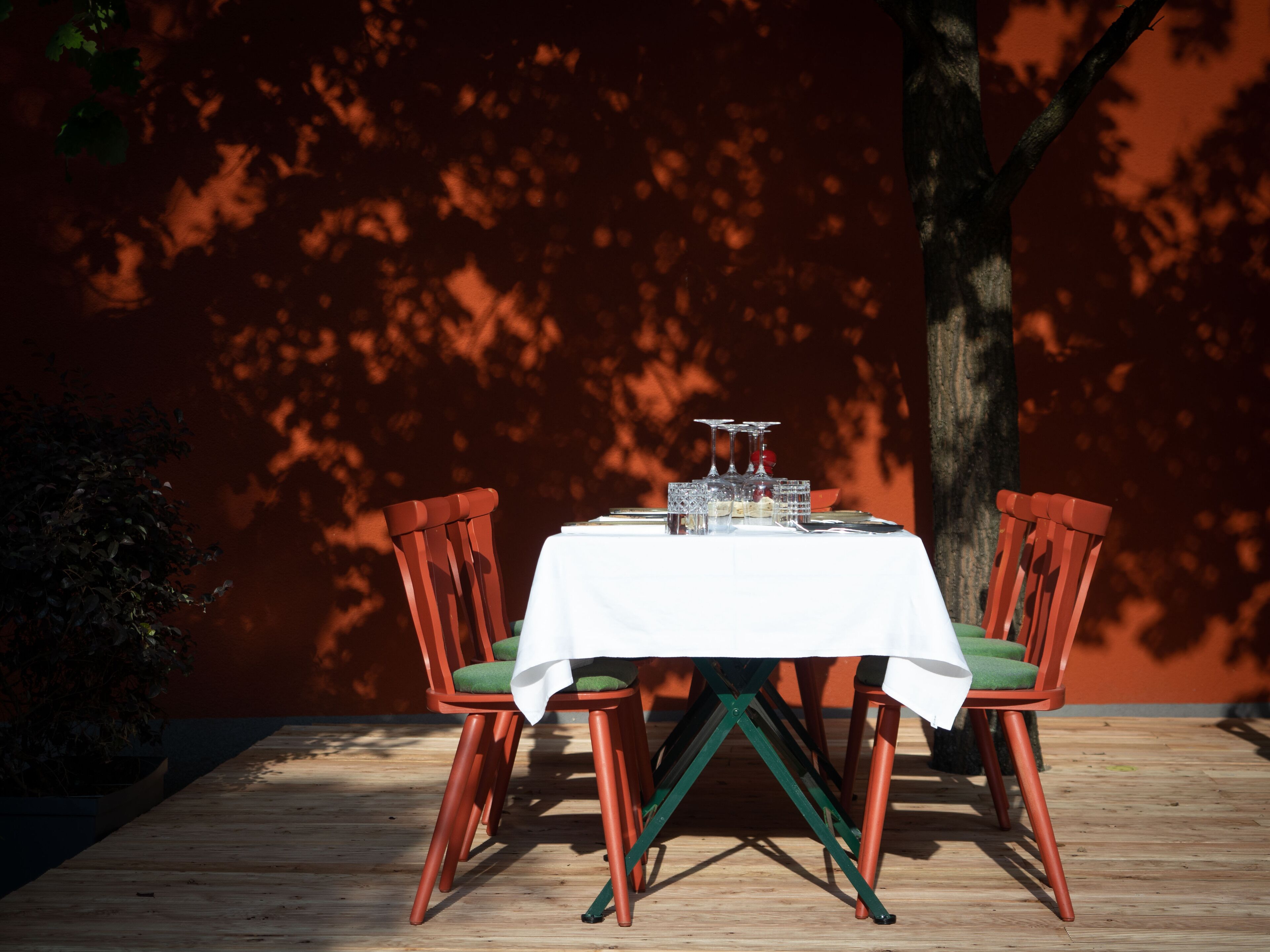 A table set outdoors with a white tablecloth, surrounded by red chairs with green cushions, against a backdrop of a tree and a red wall.