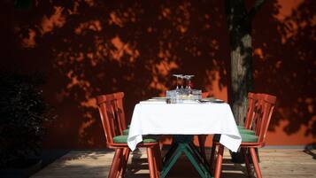 A table set outdoors with a white tablecloth, surrounded by red chairs with green cushions, against a backdrop of a tree and a red wall.