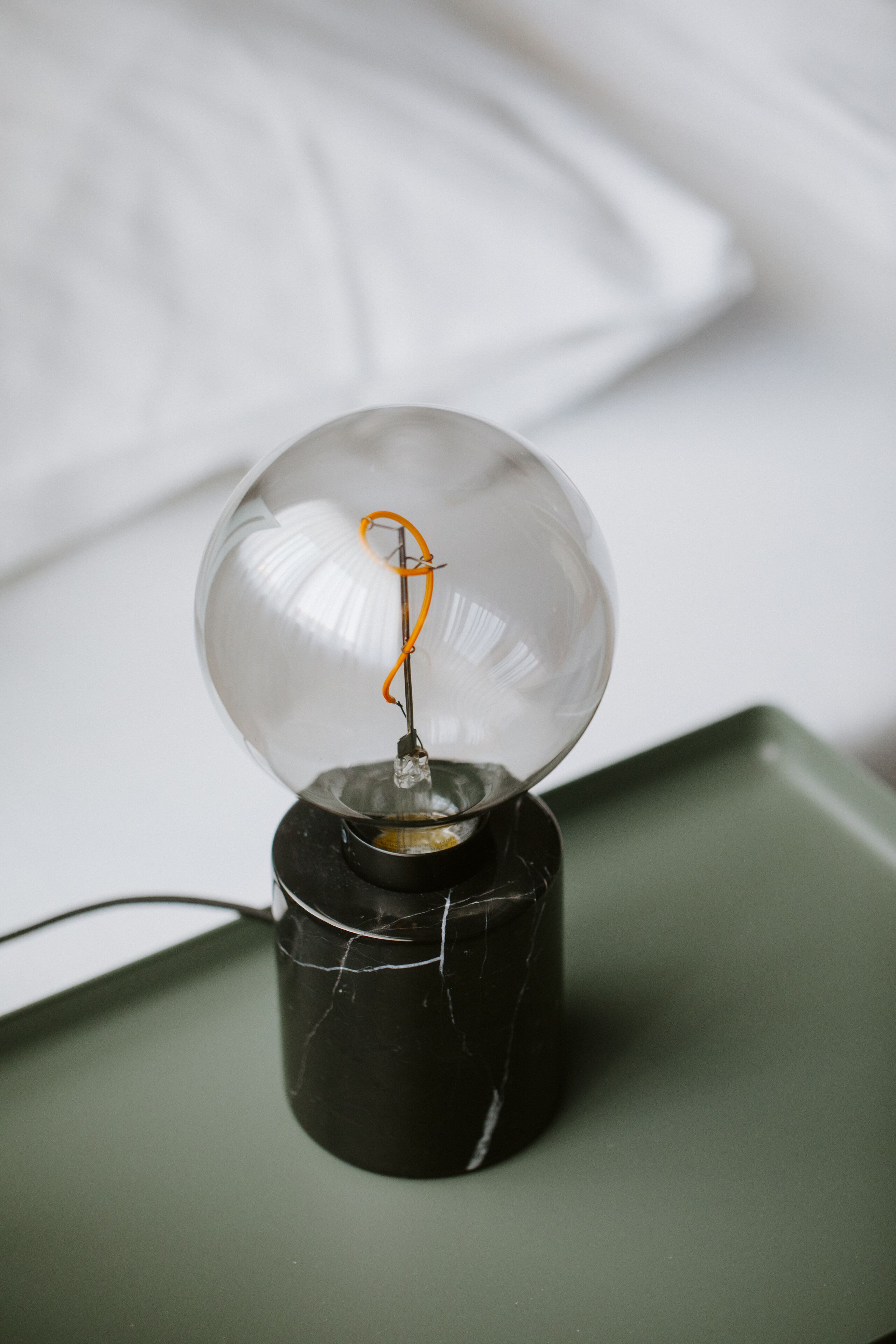 Una lampadina in vetro trasparente con un filo interno, posata su una base cilindrica nera, su una superficie verde.