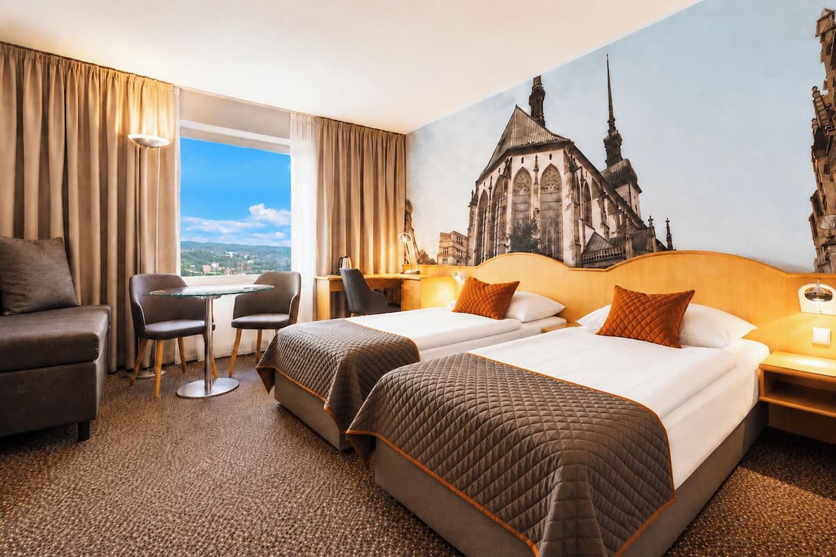 A hotel room with two beds, a desk, a chair, and a view of a cityscape.