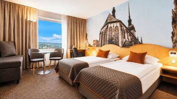 A hotel room with two beds, a desk, a chair, and a view of a cityscape.