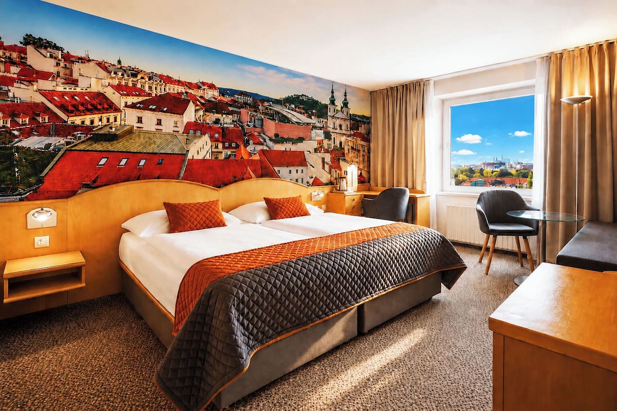 A hotel room with a bed, a desk, a chair, a window with a view of a cityscape, and a mural on the wall.