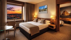 A hotel room with a large bed, two bedside tables, a desk, and a chair. There is a window with a view of a cityscape at sunset. - Cosmopolitan Bobycentrum – Czech Leading Hotels (Brno)