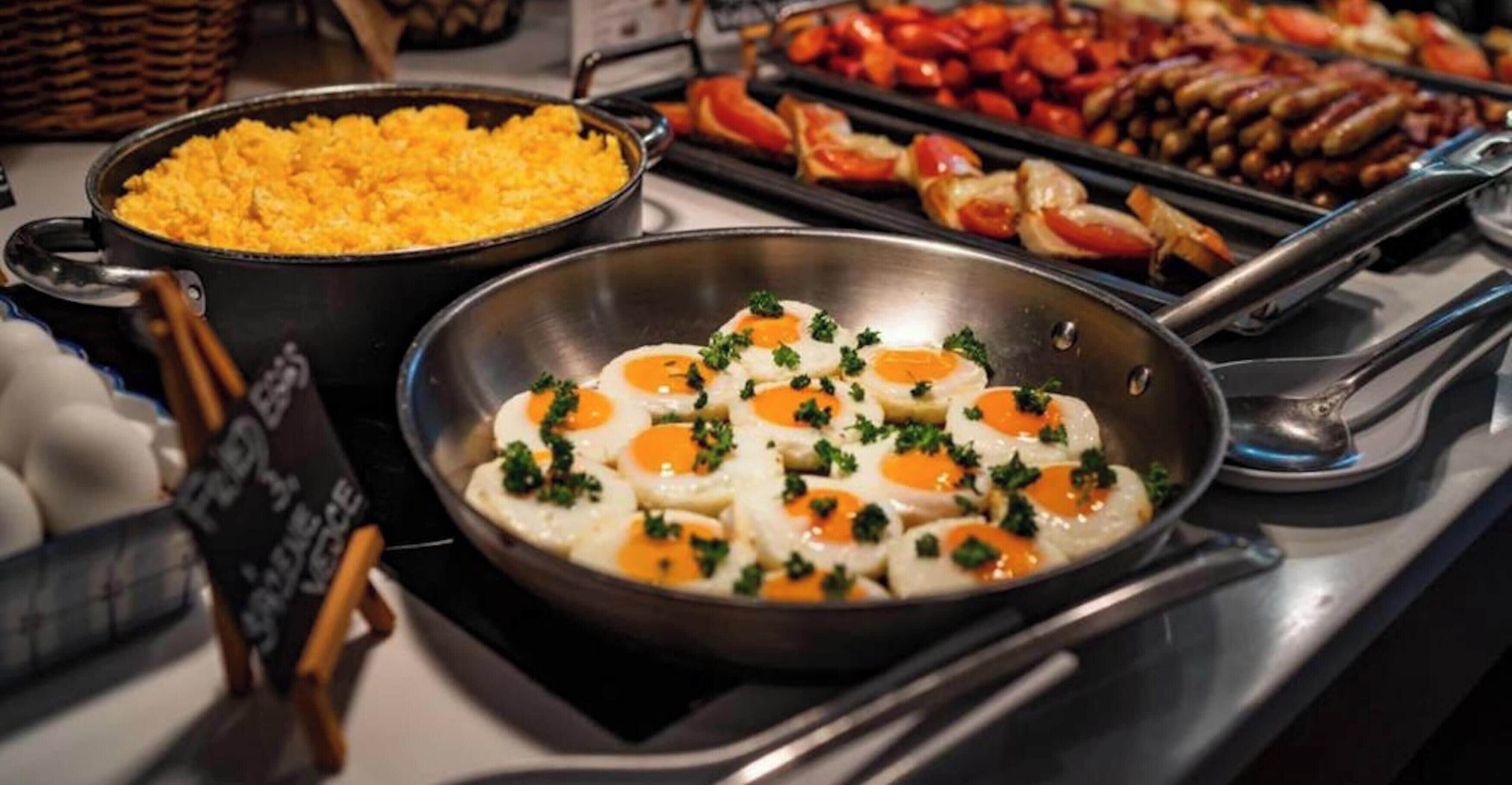 Un buffet proposant divers plats, notamment des œufs brouillés garnis d’herbes, un plat de riz et plusieurs plateaux de viandes et de légumes cuits.