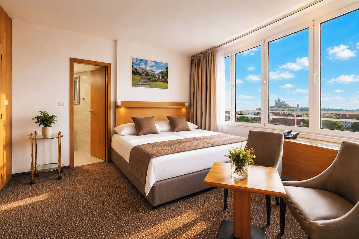 A hotel room with a bed, a desk, a chair, a view of a cityscape, and a door leading to another room.