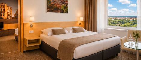 A hotel room with a large bed, bedside tables, a desk with a chair, a window with a view, and a framed picture on the wall.