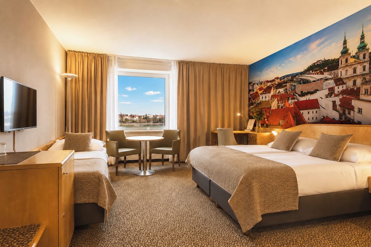 A hotel room with a large bed, a desk, a chair, a TV, and a window with a cityscape view.