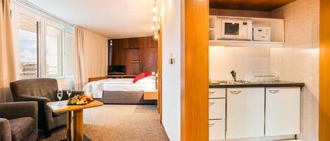 Habitación de hotel con cama, un pequeño rincón de descanso con mesa y sillas, kitchenette con microondas y lavamanos, y ventana con cortinas.