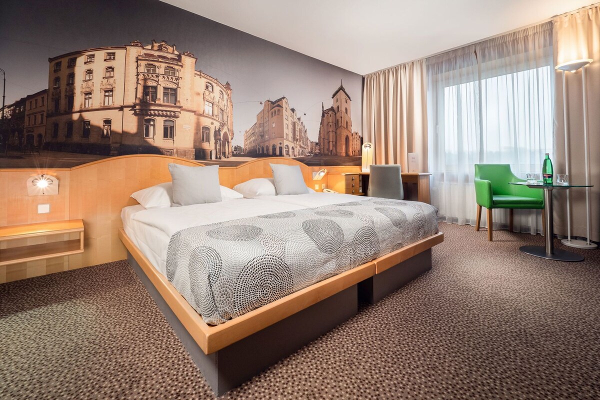 A hotel room with a large bed, a desk, a chair, a lamp, and a mural of a cityscape on the wall.
