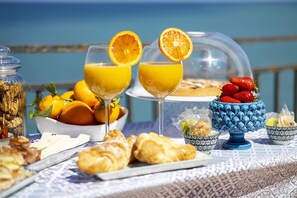 A breakfast setting with croissants, orange juice, and fruit on a table with a view of the sea. - Dolce Vita Rooms and Apartments  (Cefalù)