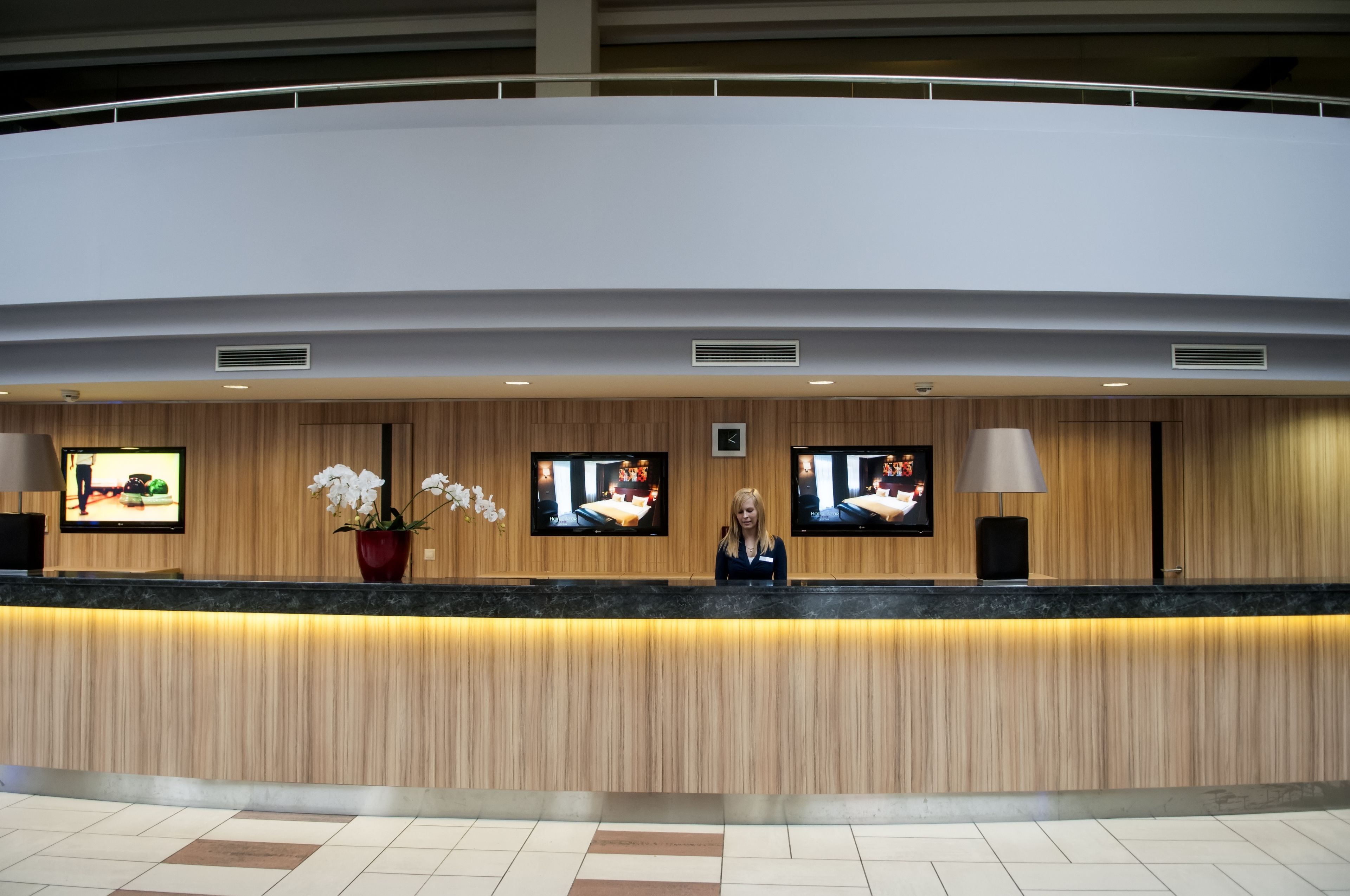 A hotel reception desk with a wooden finish, three flat-screen TVs, a potted orchid, and a lamp.