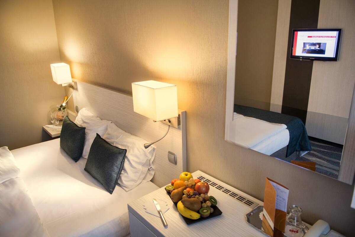 Quarto de hotel com cama bem arrumada, mesa de cabeceira com um cesto de frutas, luminária e TV fixada na parede.