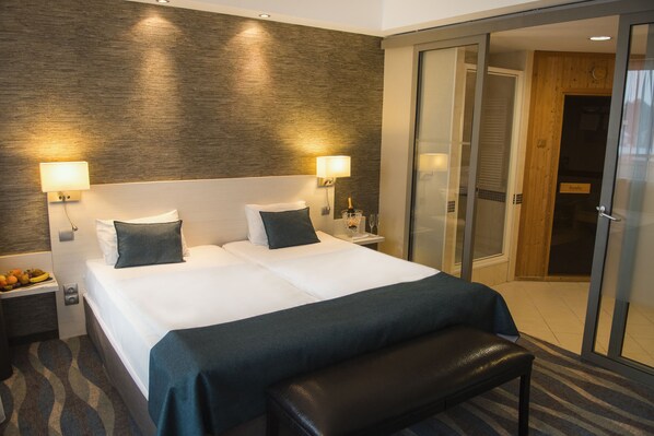 A hotel room with a large bed, two bedside tables, a bench, and a sliding glass door to a bathroom.