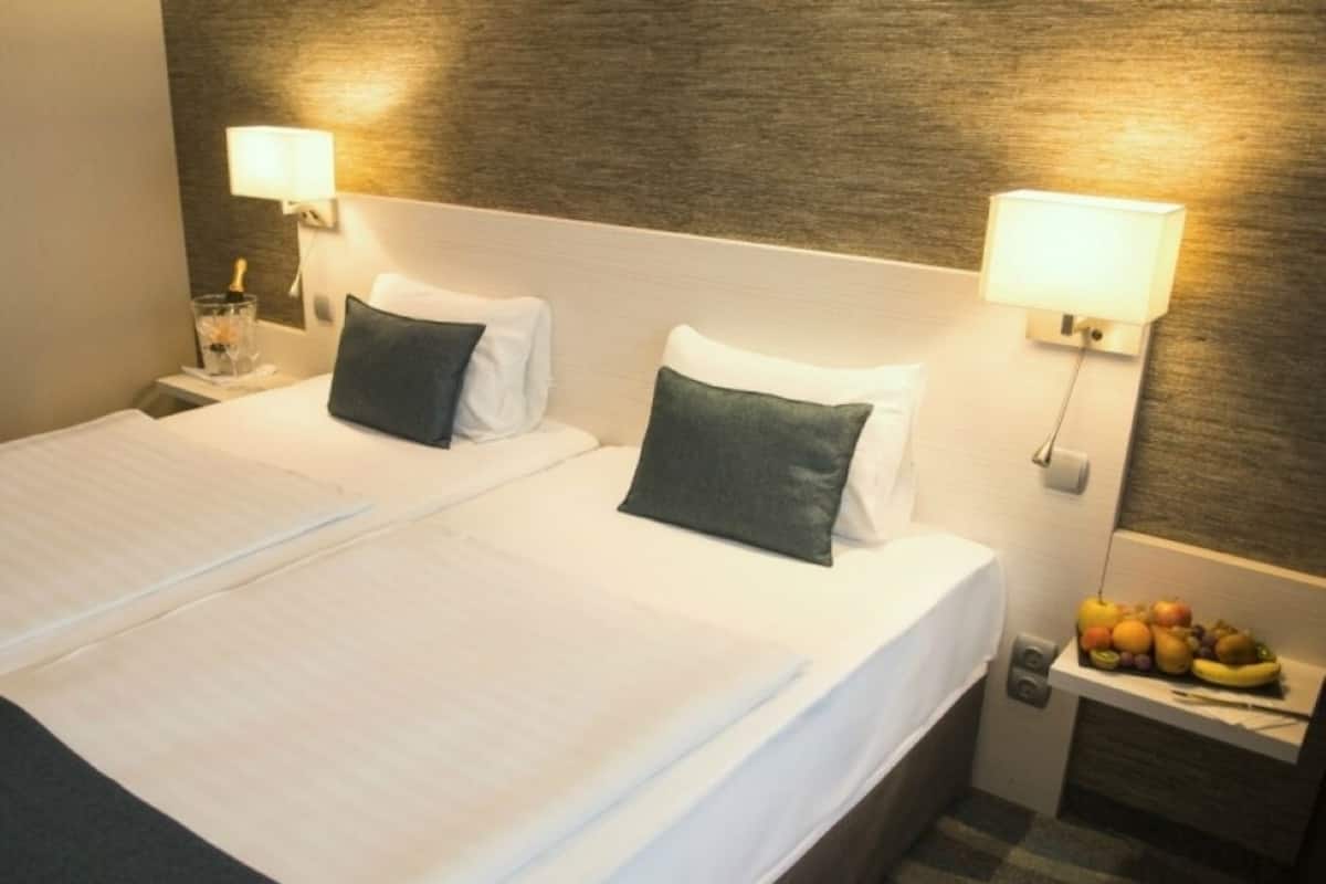 Quarto de hotel com duas camas, cada uma com lençóis brancos e almofadas escuras. Existem abajures de cada lado das camas. Uma pequena mesa com frutas é visível no canto.
