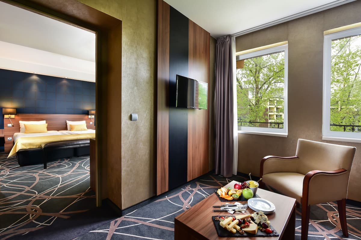Quarto de hotel com cama, televisão na parede, mesa de jantar com fruta e queijo, e vista para árvores pela janela.