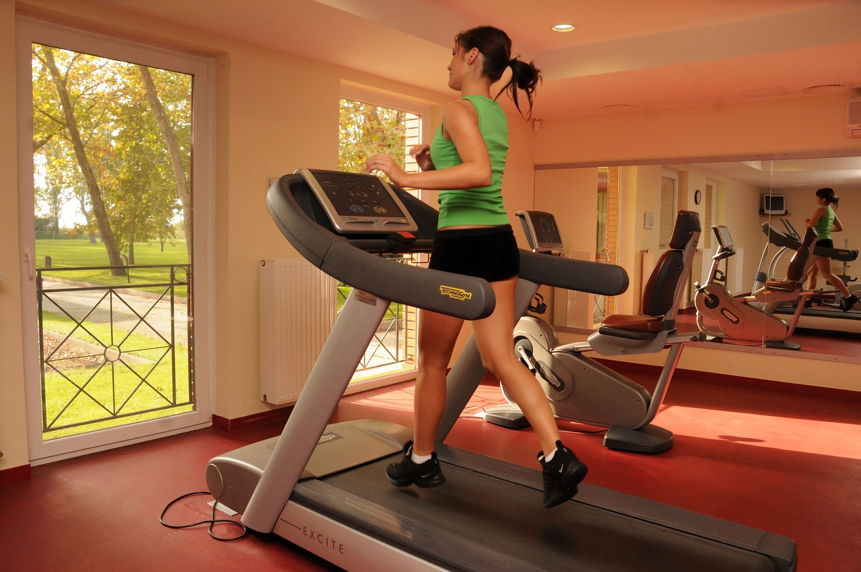 Una mujer corre en una cinta de correr en un gimnasio, con vistas a un parque a través de un amplio ventanal.