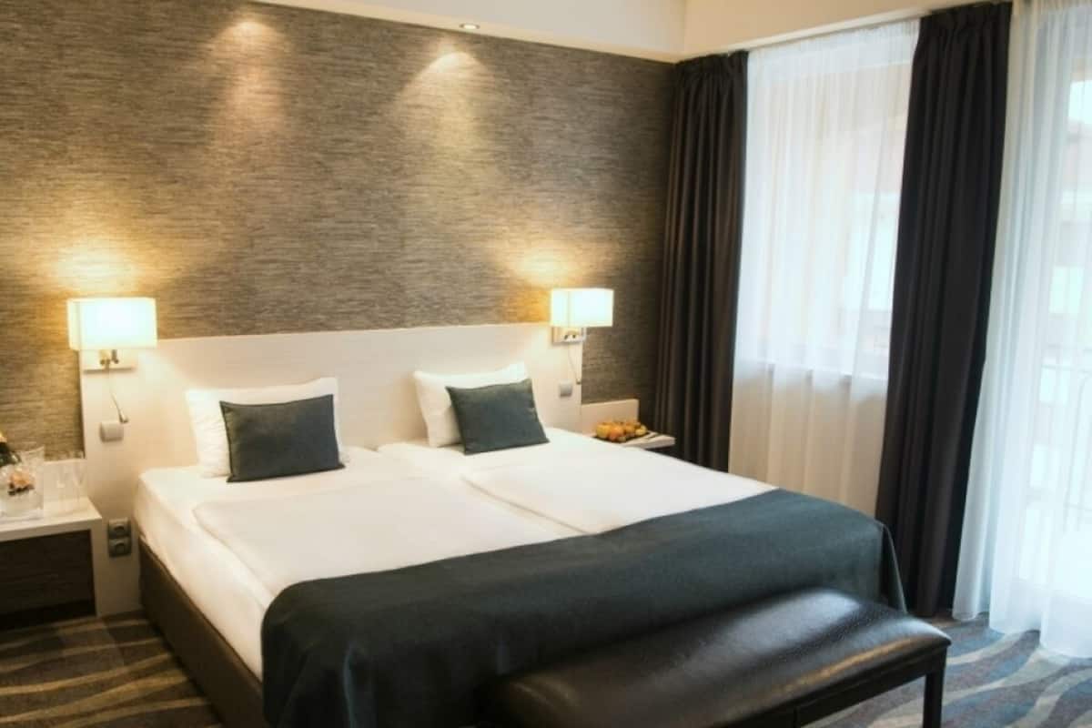 Quarto de hotel com uma cama grande, duas luminárias de cabeceira, um banquinho e vista para o exterior através de cortinas translúcidas.